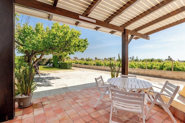 An outdoor patio with a round table and white chairs, surrounded by potted plants and a pergola structure.