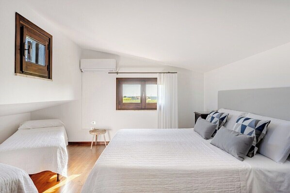 Een slaapkamer met een groot bed, een kleiner bed, een raam met gordijnen, een wandairconditioner en een kleine tafel.