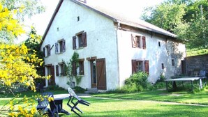 Un bâtiment à deux étages, avec une façade blanche et des volets bruns, une pelouse verdoyante, et une table et des chaises au premier plan.