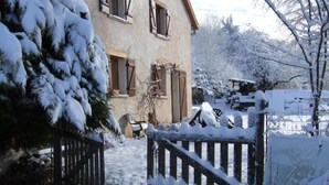 Une maison recouverte de neige, avec une porte en bois, un banc et un arbre au premier plan.