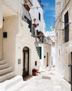 A narrow street with white buildings, a visible door, and a small step leading to a balcony with potted plants.