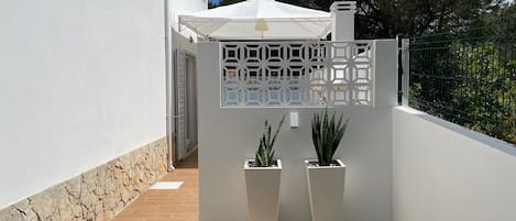 A modern outdoor patio with a white wall, stone base, two potted plants, a white canopy, and a metal fence.