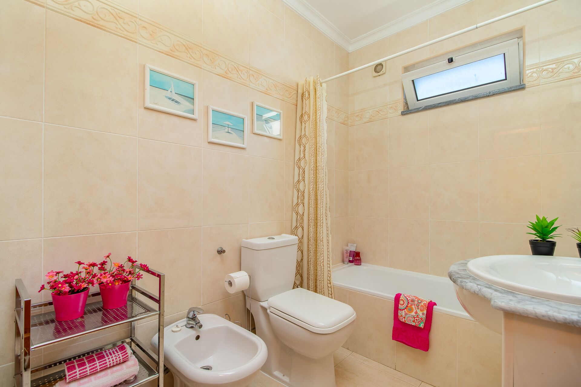 Een badkamer met een toilet, badkuip, wastafel en bidet. Er hangen foto's in lijsten aan de muur, een douchegordijn en een raam met uitzicht.