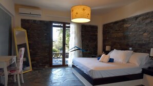 A bedroom with a stone wall, a bed with white linens, a hanging lamp, and a small table with a chair. - Family house with a sea view for rent in the area of Koundouros (Kea)