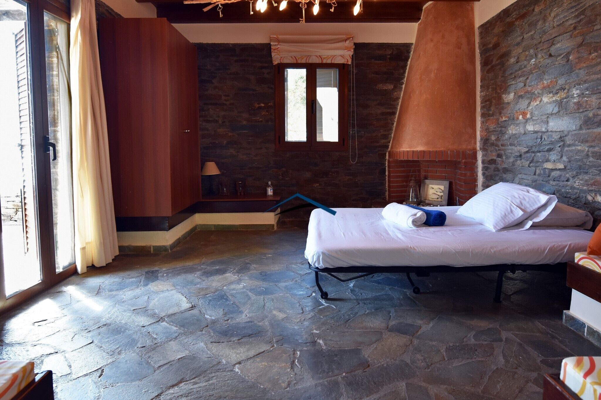 A bedroom with a stone floor, a bed with white linens, a brick fireplace, and a window with curtains.