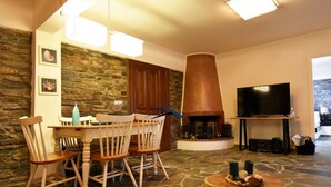 A cozy living room with a stone fireplace, a dining area with wooden chairs, a television, and a stone floor. - Family house with a sea view for rent in the area of Koundouros (Kea)