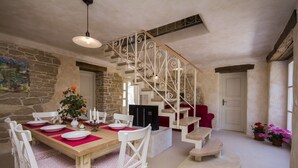 Un comedor con una mesa de madera dispuesta para cuatro personas, sillas blancas, una escalera con barandillas ornamentales y paredes de piedra.