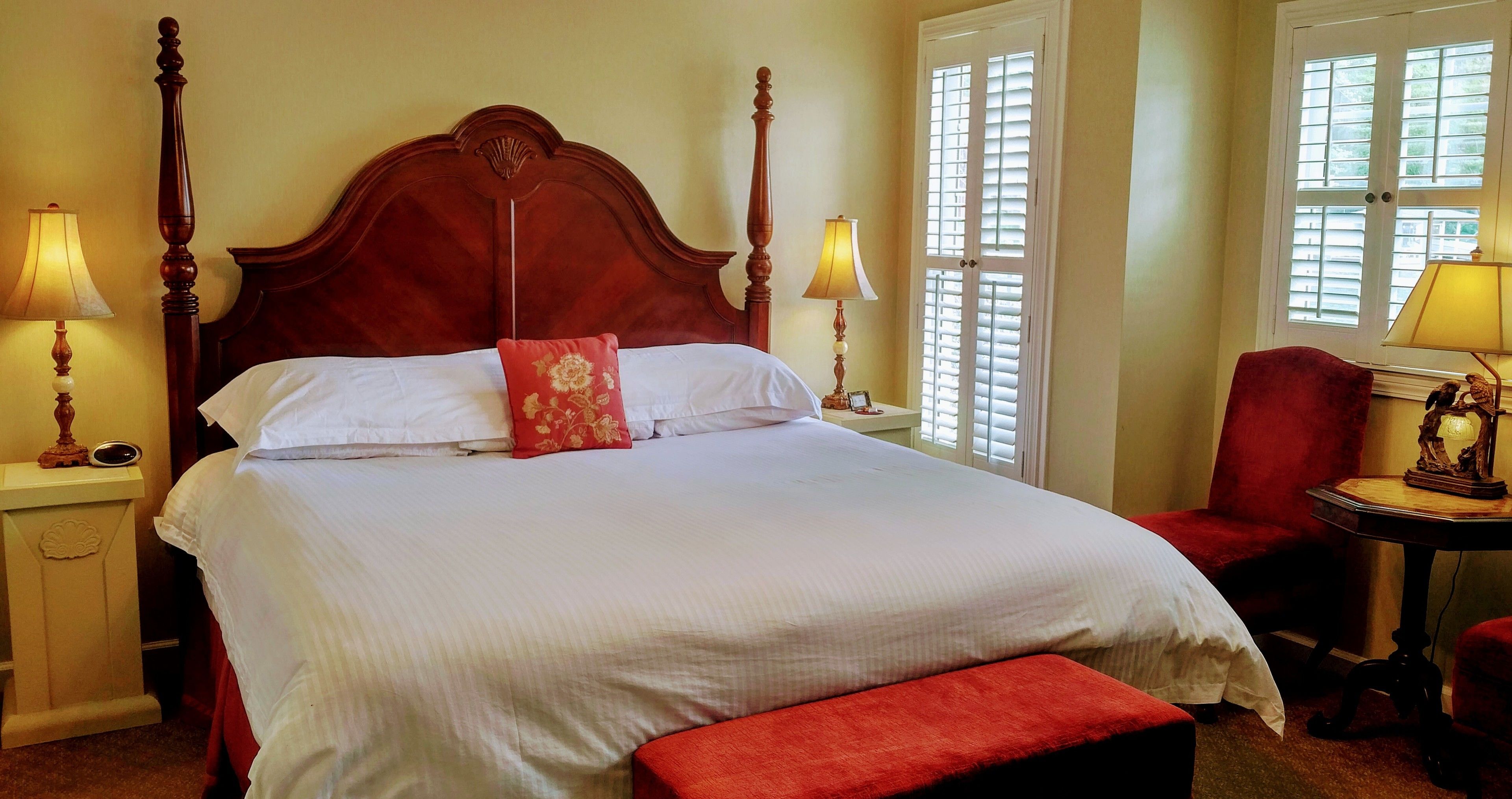 A neatly made bed with a wooden headboard, two bedside lamps, a red chair, and a small table with a lamp.