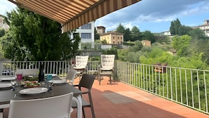 Un espace de restauration en plein air doté d’une terrasse couverte, de tables, de chaises et offrant une vue sur les bâtiments et la végétation environnants.