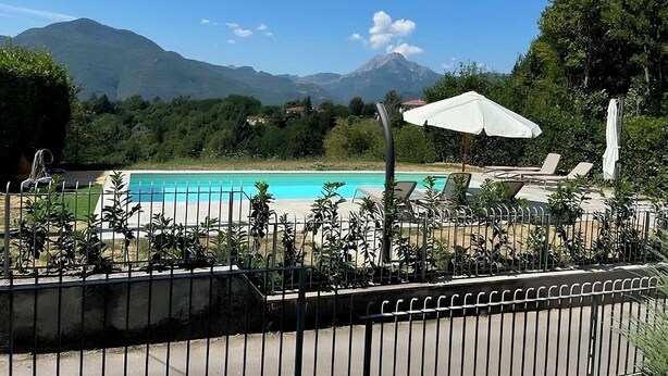 Un espace piscine doté de chaises longues et de parasols, entouré d’une clôture et de verdure, avec des montagnes en arrière-plan.