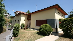 Une maison à deux étages avec un toit rouge, une grande fenêtre et un petit jardin agrémenté d’un arbuste taillé.