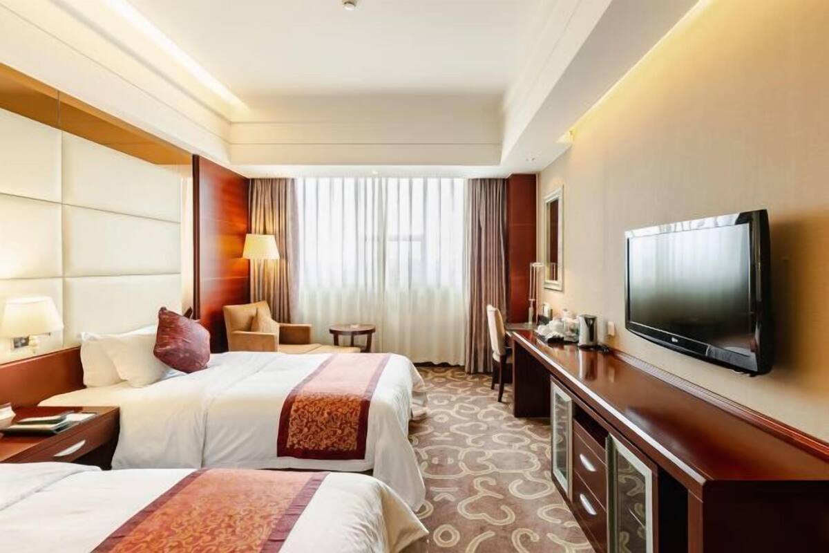 A hotel room with two beds, a desk, a television, and a large window with curtains.
