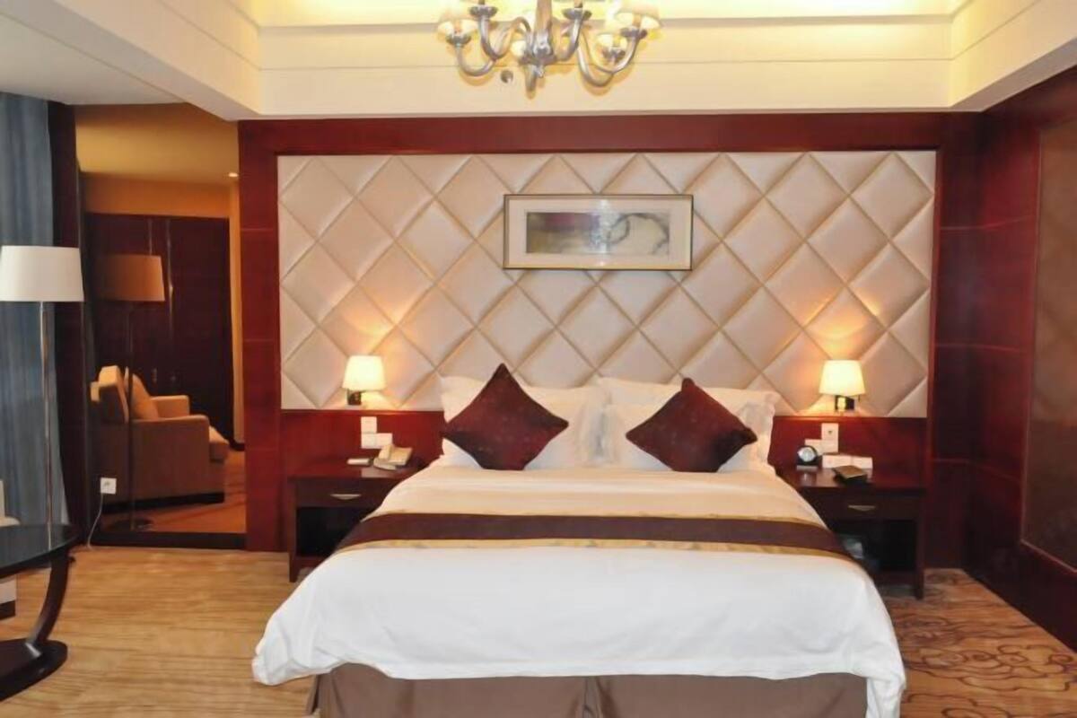 A hotel room with a large bed, two bedside tables, a chandelier, a wall-mounted artwork, and a seating area with a chair and lamp.