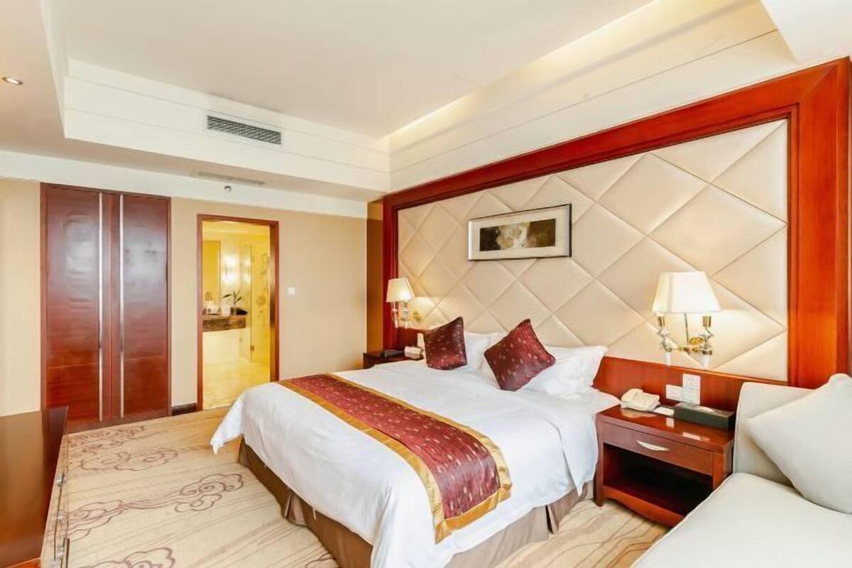 A hotel room with a large bed, a wooden headboard, a nightstand with a lamp, and a carpeted floor.