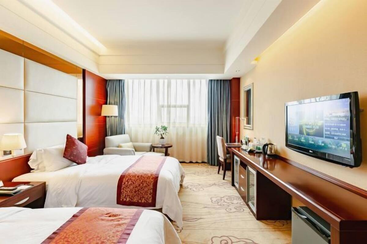 A hotel room with two beds, a desk, a TV, and a large window with curtains.