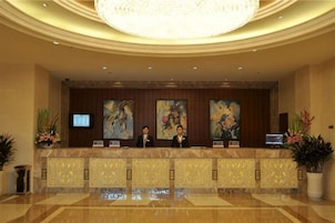 Zona de recepción de hotel con mostrador, dos recepcionistas, una lámpara de araña grande y obras de arte en la pared.