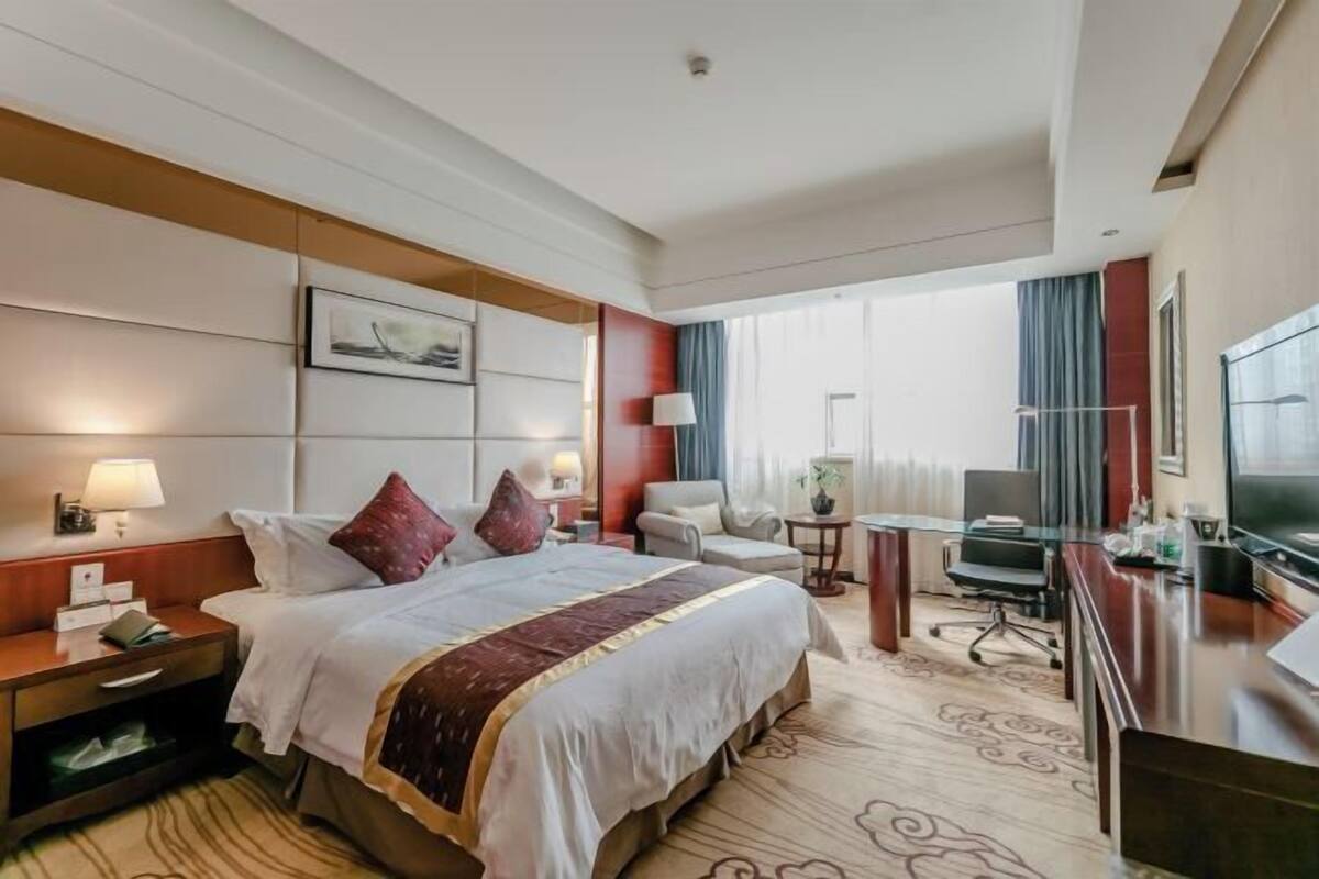 A hotel room with a bed, bedside tables, a desk, a chair, a TV, and a window with curtains.
