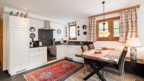 A modern kitchen with white cabinets, a black countertop, and a red patterned rug. There is a dining area with a glass table and brown chairs. - Chalet Ulysse Zermatt 4 guests /cosy/private garden/beautiful (Zermatt)