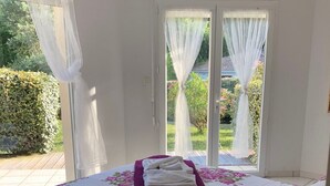 Un dormitorio con cama, ventana con cortinas y vistas al exterior con vegetación, además de una lámpara sobre una mesita de noche.