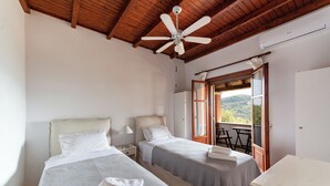 A bedroom with two beds, a ceiling fan, a window with a view, and a table by the window. - Stathis House - Ag. Georgios Corfu (Pagi)