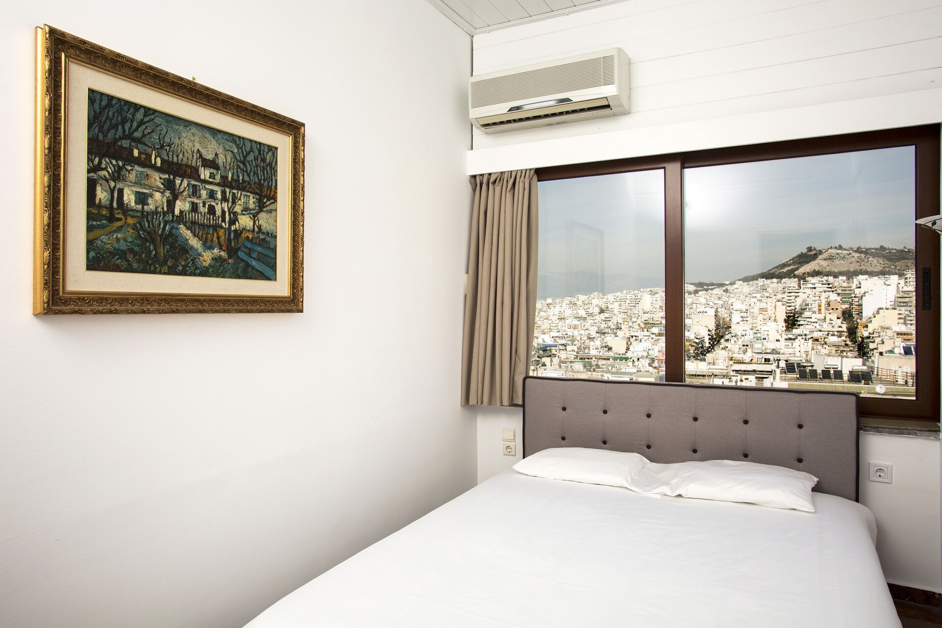 A hotel room with a bed, a large window with a city view, an air conditioner, and a framed painting on the wall.