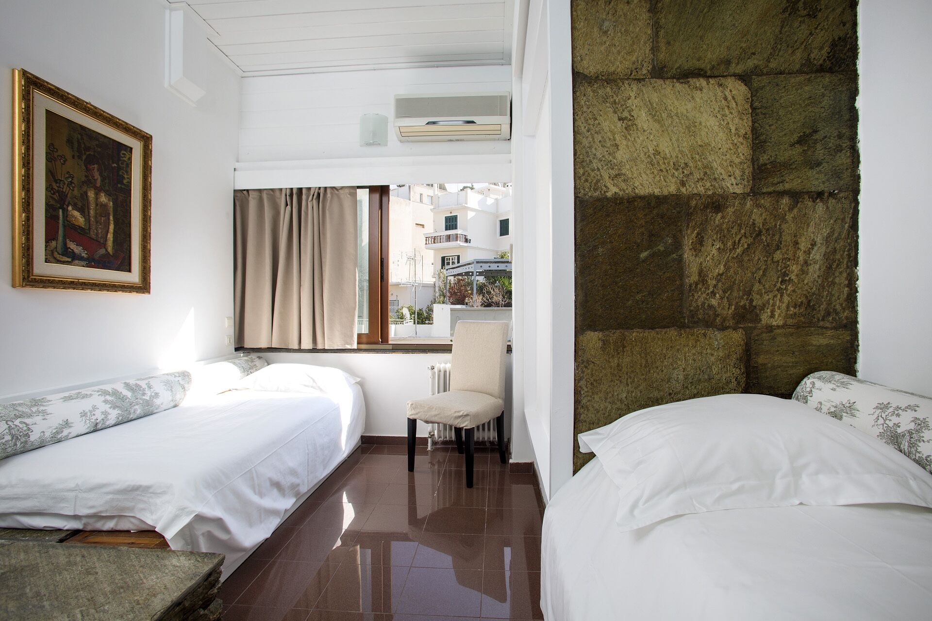 A hotel room with two beds, a painting on the wall, a window with curtains, and a view of buildings outside.