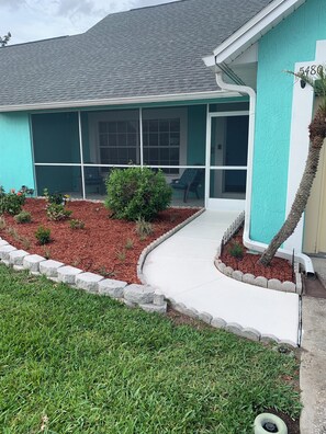 A house with a green exterior, a covered porch, and a well-maintained front yard with a stone border and mulch.