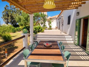 Eine Terrasse im Freien mit einem Holztisch und -stühlen, einer hängenden Leuchte und Blick auf ein weißes Gebäude mit einem Ziegeldach.