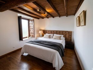 A bedroom with a wooden ceiling, a bed with white bedding, a nightstand with a lamp, and a window with a view.