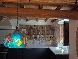 A kitchen with a hanging colorful light fixture, wooden beams, and a stone wall backsplash.