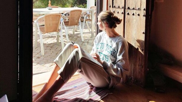 Una persona sentada en el suelo leyendo un libro en una habitación con una puerta de madera, una ventana y vistas a un patio exterior con sillas y una mesa.