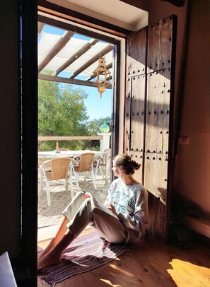 Une personne est assise sur le sol, en train de lire un livre dans une pièce dotée d’une porte en bois, d’une fenêtre et donnant sur une terrasse extérieure avec des chaises et une table.