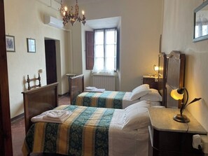 Habitación de hotel con dos camas, un escritorio, una lámpara, un espejo, un ventanal con cortinas, un candelabro y un radiador.