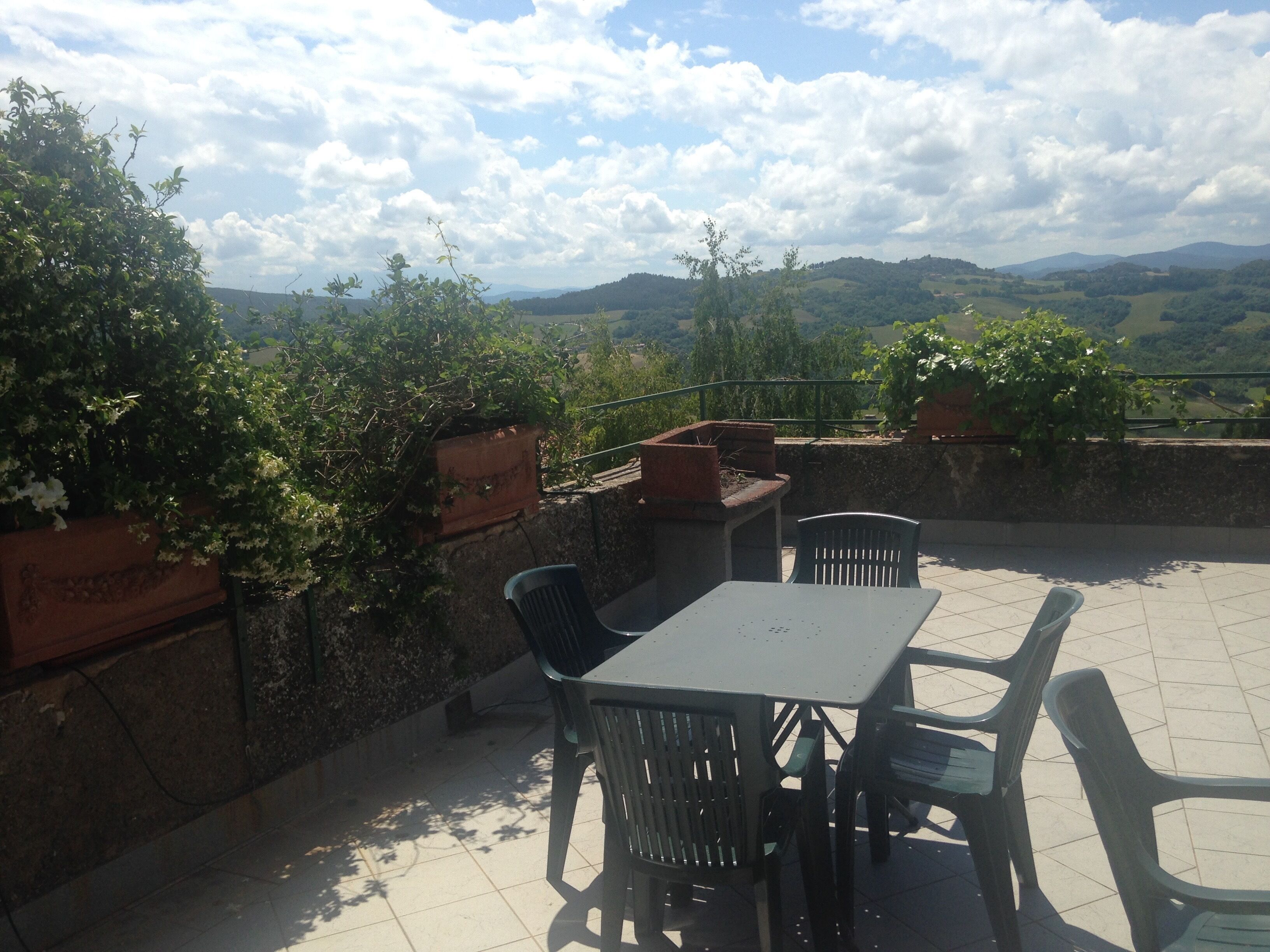En udendørs terrasse med et bord og stole, omgivet af plantekasser og med udsigt over et smukt landskab.
