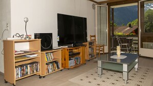 A living room with a TV, bookshelves, a coffee table, and a view of mountains through a sliding glass door. - 2 BALCONIES,1 TERRACE,VIEWS OF  WATERFALL, MOUNTAINS, RIVER,WENGEN,TRAINS, (Lauterbrunnen)