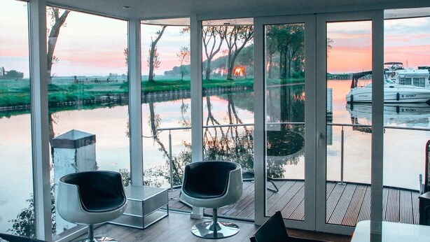 Une chambre moderne dotée de grandes fenêtres donnant sur une rivière paisible et des bateaux, et comprenant deux fauteuils contemporains et une table.
