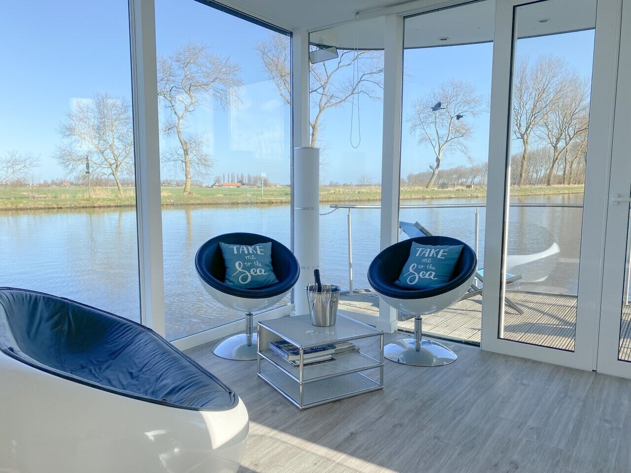A modern room with large windows overlooking a body of water, two black chairs with cushions, a small table with a book and a drink, and a white chair.