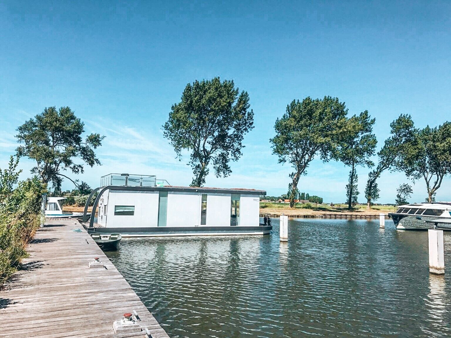Ein Hausboot liegt an einem hölzernen Steg, im Hintergrund fließt ein ruhiger Fluss und Bäume.