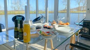 Une table à manger avec de la nourriture et des boissons, de grandes fenêtres donnant sur l’eau et les arbres, et une cloison en verre.