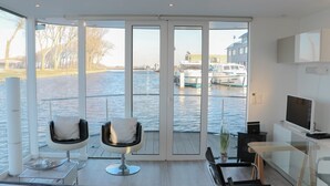 Une chambre moderne dotée d’une grande porte-fenêtre coulissante donnant sur une vue sur l’eau, de deux fauteuils, d’une petite table, d’un téléviseur et d’une plante.