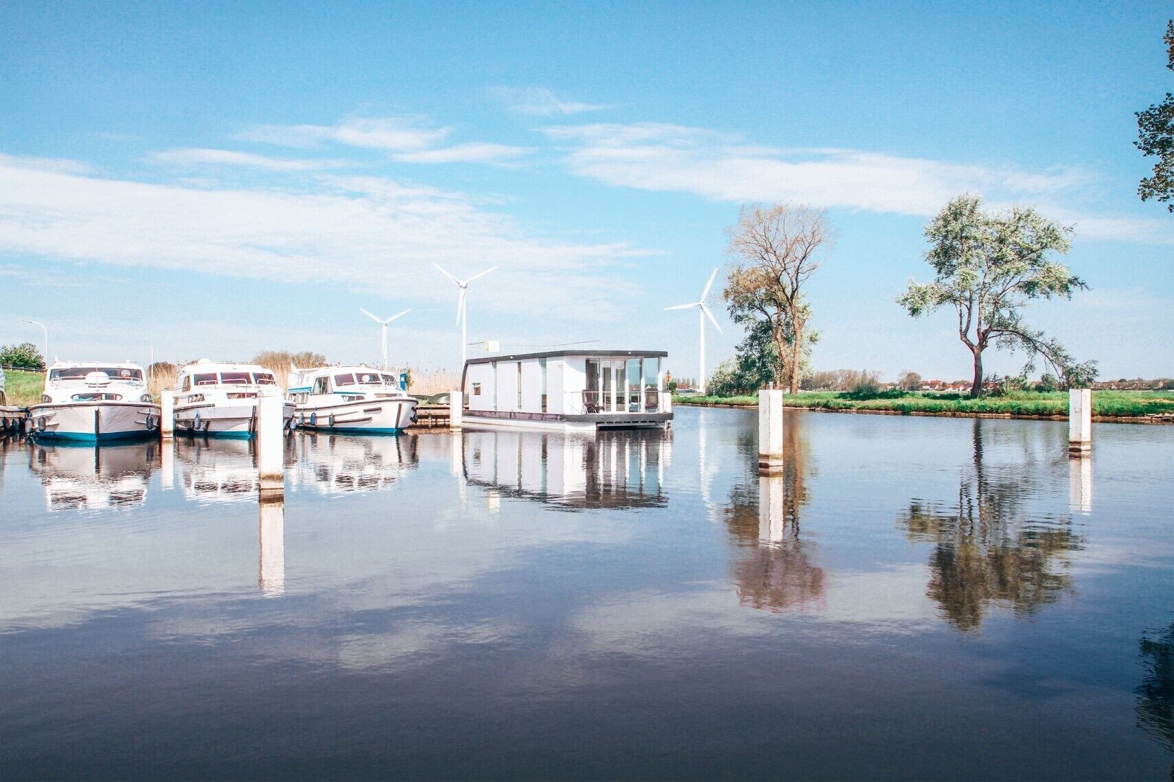 A calm water body with boats docked, a modern building, and wind turbines in the background.