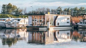 Une péniche flottante est amarrée à une marina où se trouvent plusieurs bateaux, le tout étant entouré d’arbres et surplombé par un ciel dégagé.