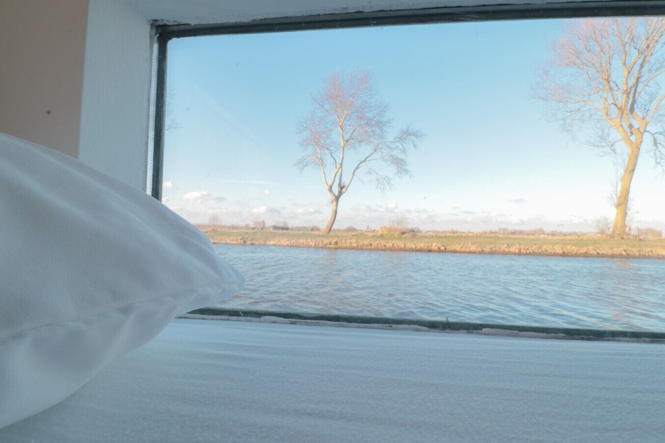 Un lit avec un oreiller blanc et une fenêtre donnant sur une rivière bordée d’arbres.