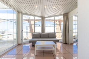A modern interior with a sofa, a coffee table, and large windows offering a view of the sky and a brick structure outside. - AirCon Penthouse Sea Views · Parking Pool · CENTER II 113 (Salou)