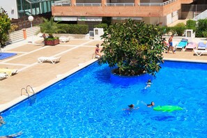 A swimming pool with a tree in the center, surrounded by lounge chairs and a building in the background. - AirCon Penthouse Sea Views · Parking Pool · CENTER II 113 (Salou)
