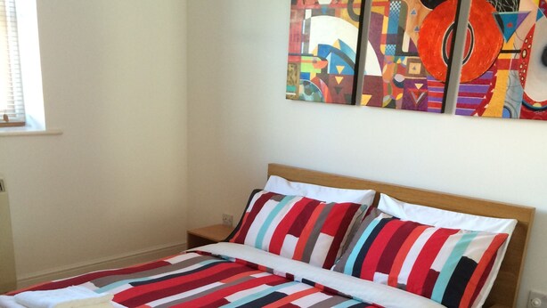 A bed with a striped bedspread and pillows, a wooden headboard, and a nightstand with a lamp. - Beautiful Kingston apartment (Kingston upon Thames)