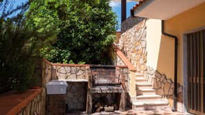 Un espace barbecue extérieur en pierre, avec un banc en pierre et un évier, entouré de verdure et agrémenté d’un escalier menant à une porte.