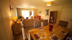A dining area with a wooden table set for four, surrounded by upholstered chairs. There is a sofa and a bookshelf in the background.
