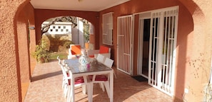 A covered patio with a table and chairs, white shutters, and a view of a building in the background.