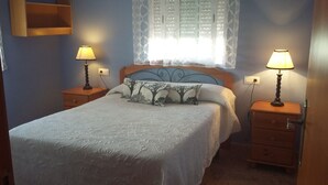 Een slaapkamer met een bed, twee nachtkastjes met lampen, een raam met gordijnen met dessin, en een wandplank.